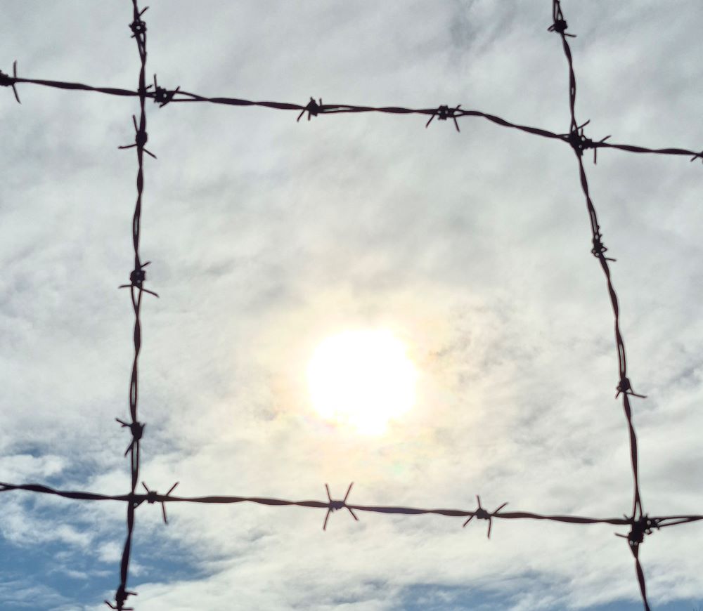 Symbolbild: Wolkiger Himmel mit etwas Sonne hinter Stacheldraht